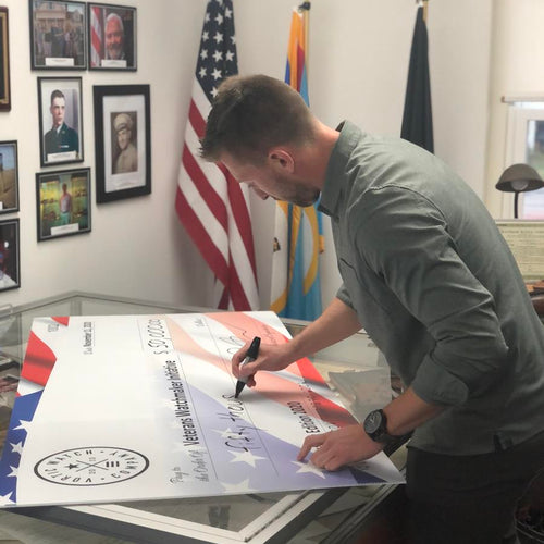 Man signing a very large check, American flag in the background. 