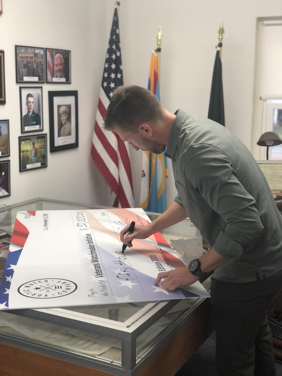 Man signing a very large check, American flag in the background. 