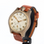 A vintage-style wristwatch with a round bronze-toned case, cream-colored dial, gold-tone numerals and hands, and a brown leather strap, shown on a plain white background.