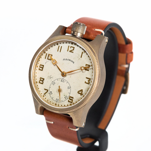 A vintage-style wristwatch with a round bronze-toned case, cream-colored dial, gold-tone numerals and hands, and a brown leather strap, shown on a plain white background.