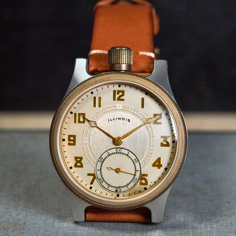 A vintage-style wristwatch with a round silver-toned case, cream-colored dial, gold-tone numerals and hands, and a brown leather strap, photographed standing upright on a textured surface against a dark blurred background.