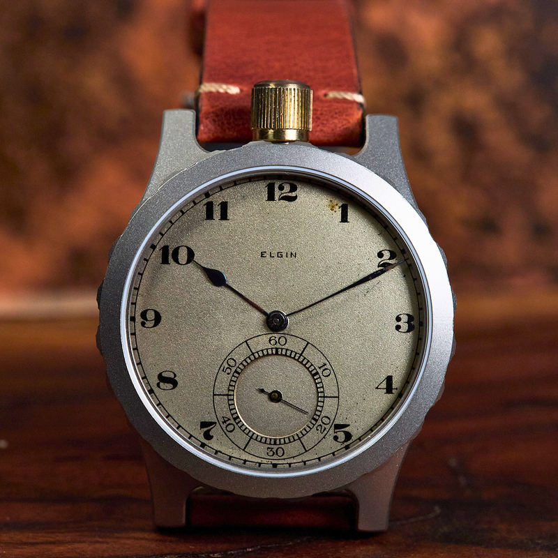A vintage-style wristwatch with a round silver-toned case, a beige dial with black Arabic numerals, and a brown leather strap, photographed against a warm brown blurred background.