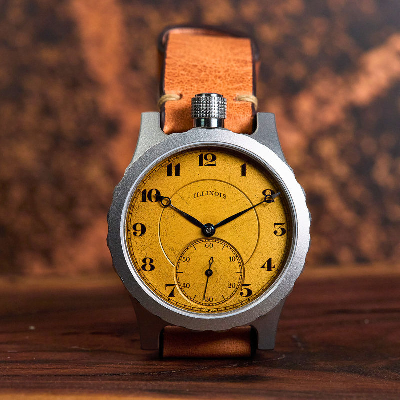 A close-up shot of a vintage-style wristwatch with a round silver-tone case, a textured yellow dial, and a brown leather strap, resting on a dark wooden surface with a blurred background.