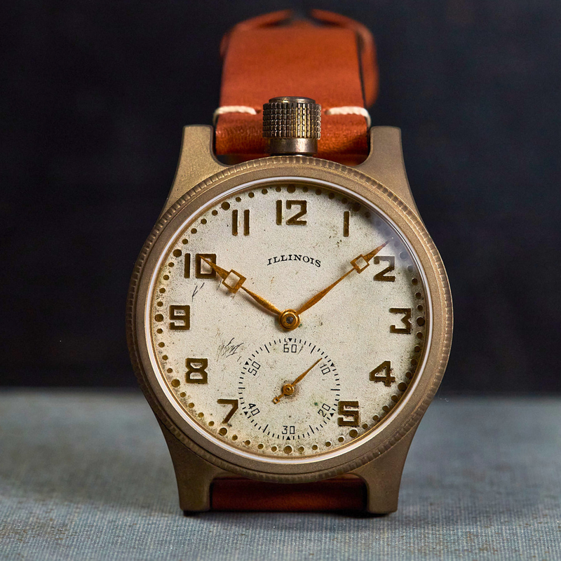 A vintage-style wristwatch with a round bronze-toned case, cream-colored dial, black Arabic numerals, and a brown leather strap, photographed against a dark background with a textured surface below.