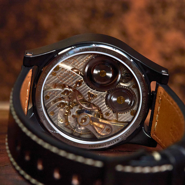 Close-up of a black smartwatch with a transparent back showing a detailed mechanical movement, attached to a brown leather strap on a wooden surface.