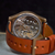 Close-up of the back of a wristwatch with a tan leather strap and a rose gold-toned case, showing a detailed exposed mechanical movement through a transparent circular backplate.