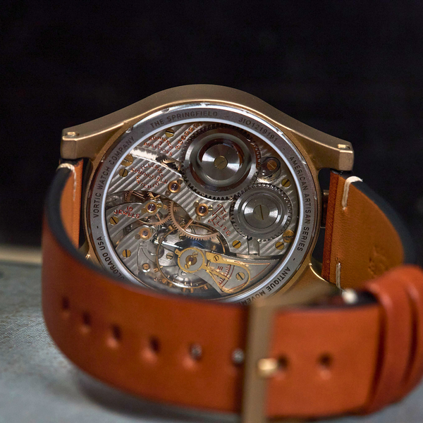 Close-up of the back of a wristwatch with a tan leather strap and a rose gold-toned case, showing a detailed exposed mechanical movement through a transparent circular backplate.