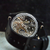 A close-up of a black leather watch strap with a transparent back showing a detailed mechanical movement with silver and gold-toned gears, set against a dark textured background.