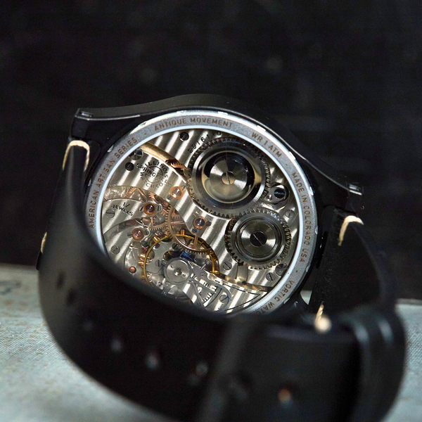 A close-up of a black leather watch strap with a transparent back showing a detailed mechanical movement with silver and gold-toned gears, set against a dark textured background.