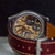 A close-up shot of a skeleton watch showing its intricate gold-toned mechanical movement through a dark brown leather strap with white stitching.