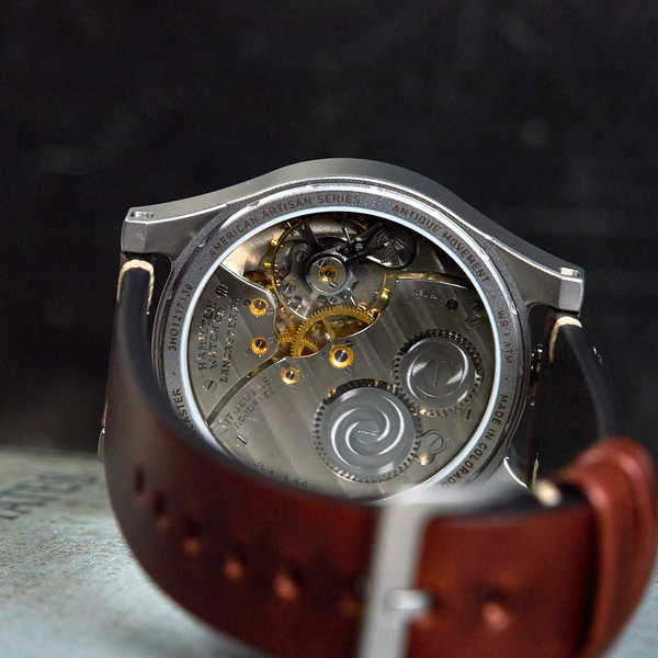 Close-up of the back of a wristwatch with a brown leather strap, showing a detailed silver-tone automatic mechanical movement through a sapphire crystal.
