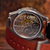 Close-up of a wristwatch with a transparent back showing a detailed mechanical movement, attached to a brown leather strap on a wooden surface.