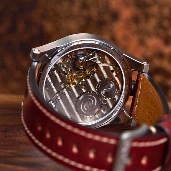 A close-up shot of a luxury watch with a silver-toned case, showing the intricate mechanical movement through a transparent backplate, attached to a dark red leather strap on a wooden surface.