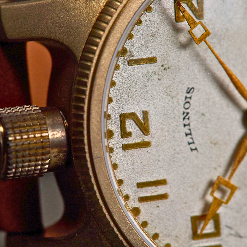 Close-up of a vintage-style watch with a gold-toned case, textured crown, off-white dial, gold hands and numerals, and a reddish-brown leather strap.