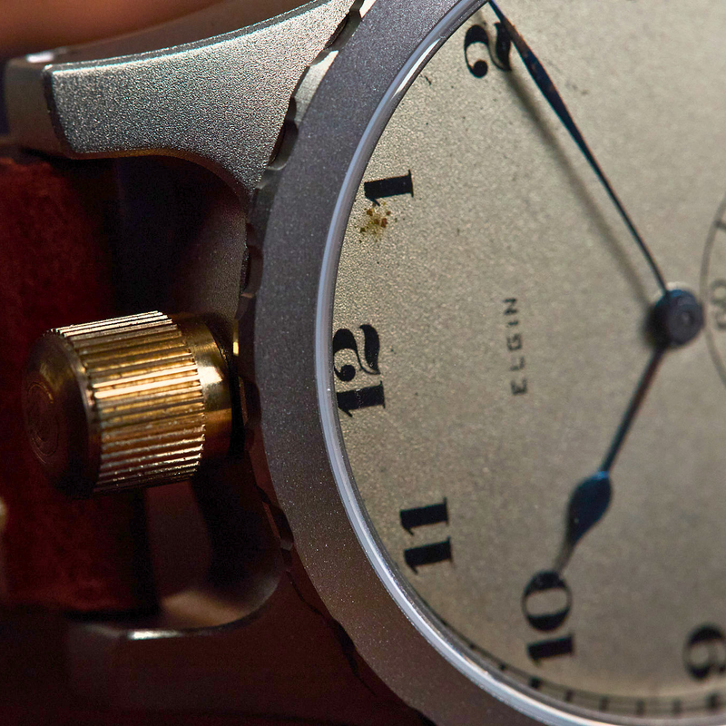 Close-up of an Elgin wristwatch with a silver-toned case, textured beige dial, black numerals, and a reddish-brown leather strap.