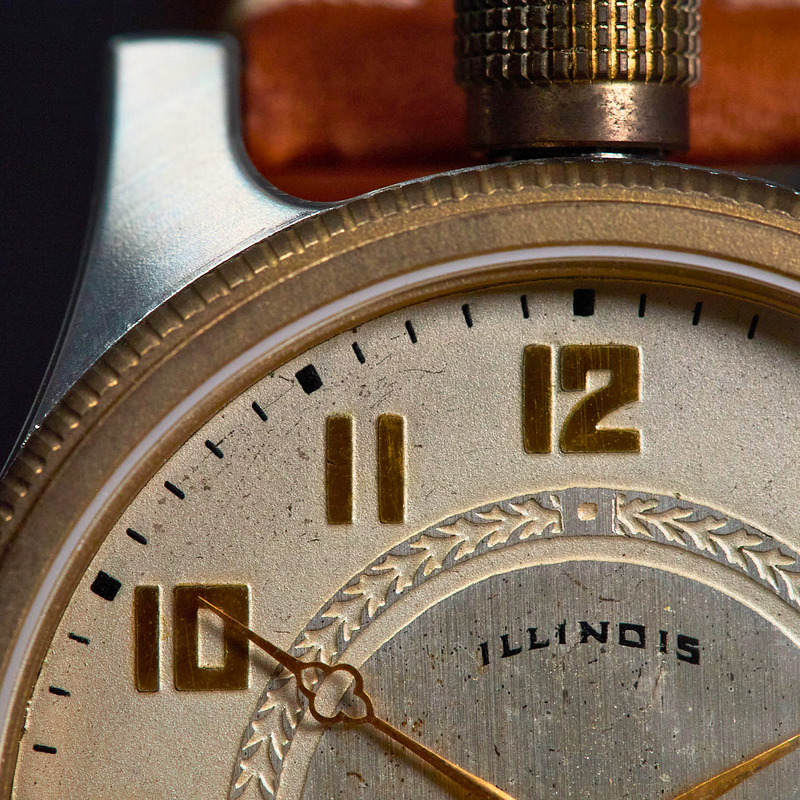Close-up of a vintage-style wristwatch face with a cream dial, gold-tone numerals and hands, and a dark textured bezel. The word 'ILLINOIS' is visible on the dial.