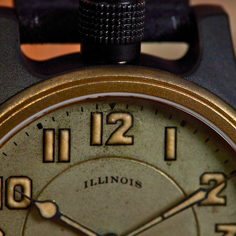 Close-up of a vintage-style watch with a black case, gold-tone bezel, and an aged cream dial featuring the word 'ILLINOIS' near the center.