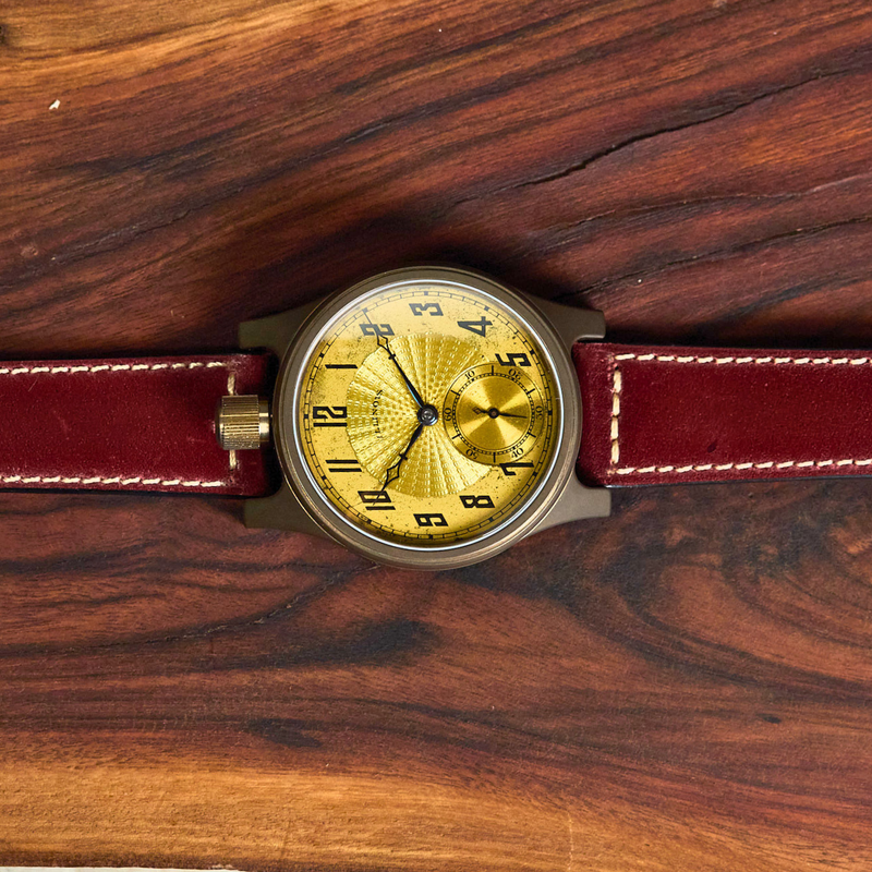 A wristwatch with a round gold dial, black numerals, and a dark brown leather strap, laid flat on a dark wood background.