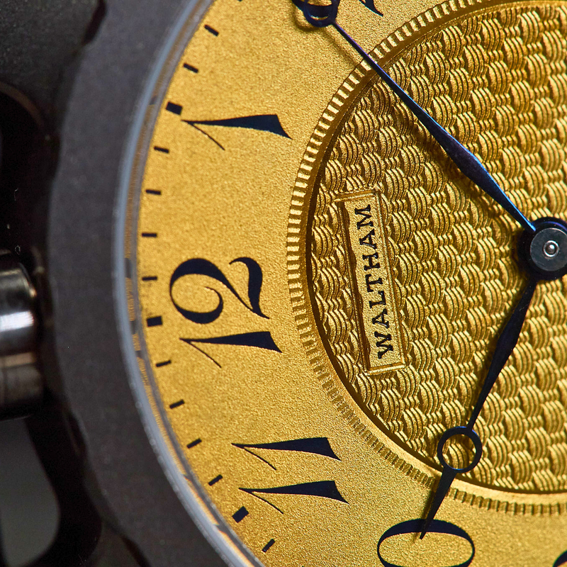 Close-up of a Waltham watch face with a textured gold dial, black Arabic numerals, black hands, and a dark gray case.