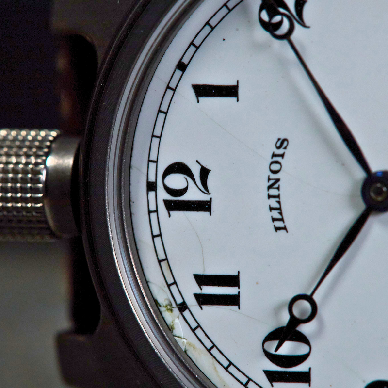 Close-up of a wristwatch face showing black Arabic numerals on a white dial with the word ILLINOIS printed below the 12, plus a silver textured crown and part of a dark case.