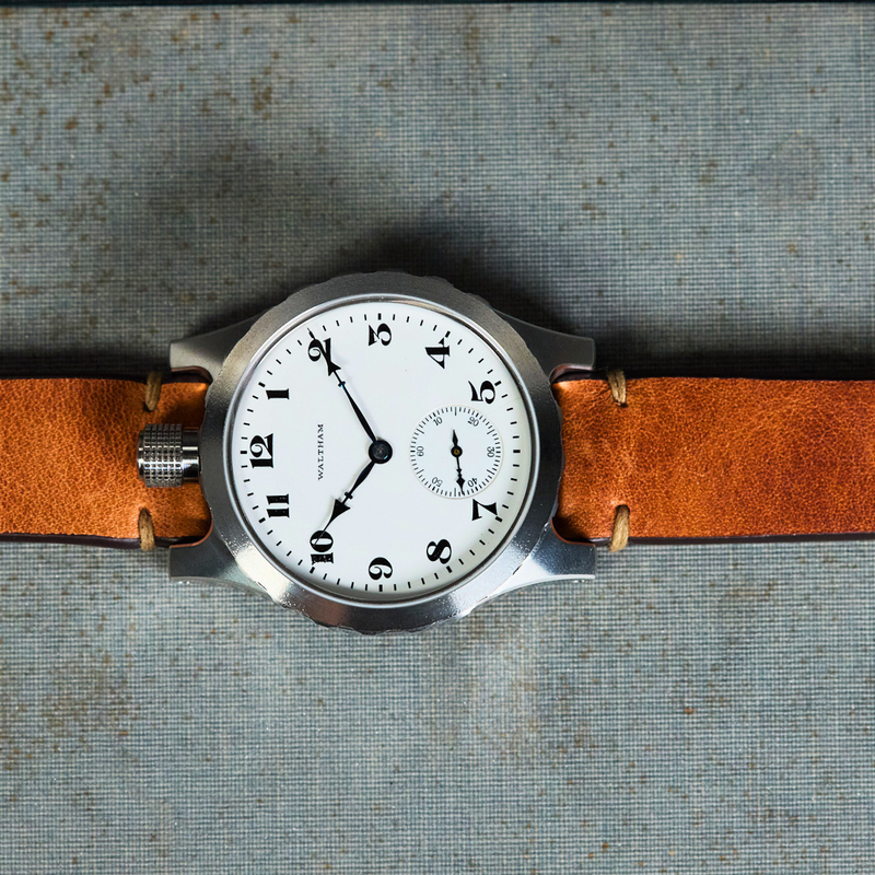 A wristwatch with a white dial, black numerals, and a silver-toned case, laid flat on a textured gray surface with a brown leather strap extending left and right. The strap is positioned over printed text on a book or magazine page.