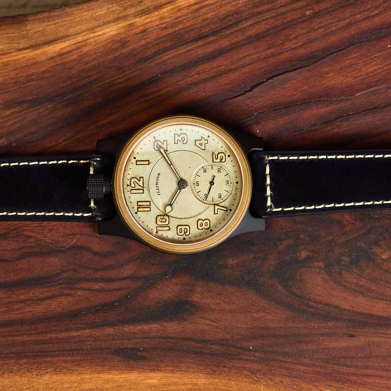 A vintage-style wristwatch with a round beige dial, black case, and black leather strap with white stitching, displayed horizontally on a dark wood surface.