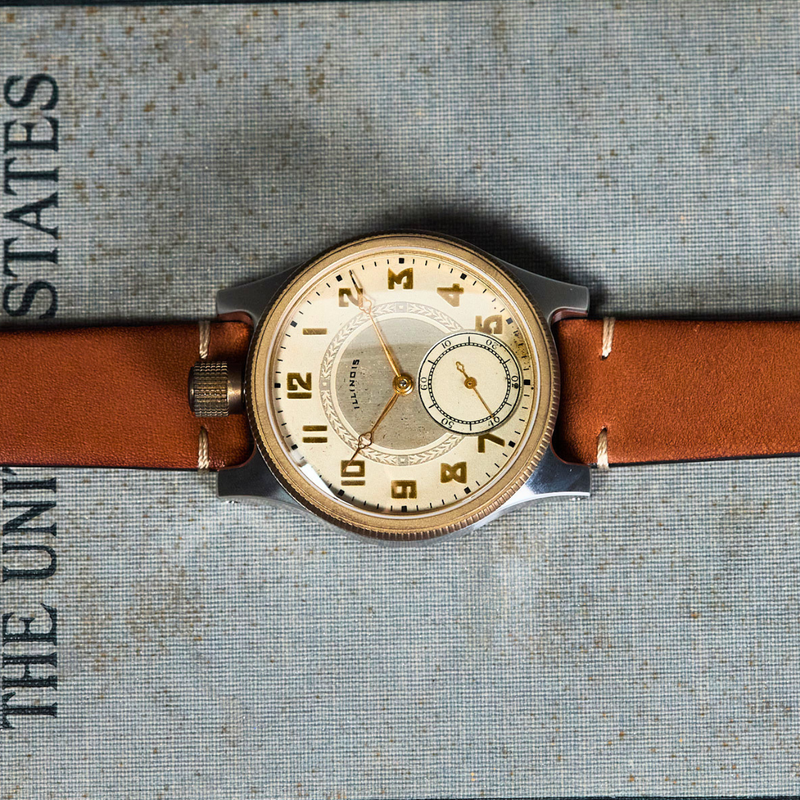 A vintage-style wristwatch with a tan leather strap and a cream-colored dial, laid flat on a textured gray surface with printed text.