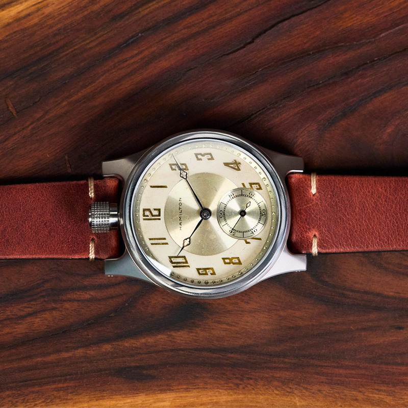 A vintage-style wristwatch with a round silver-toned case, cream dial, and reddish-brown leather strap, laid flat on a dark wood surface.