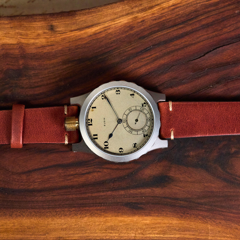 A vintage-style wristwatch with a round silver-toned case, cream dial, and reddish-brown leather strap, laid horizontally on a polished dark wood surface.