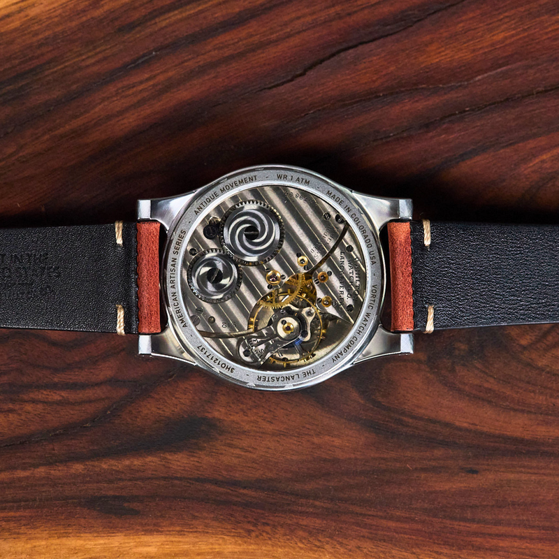 A close-up of a skeleton watch with a black leather strap and a silver-toned case showing the intricate inner mechanism, laid flat on a dark wood background.