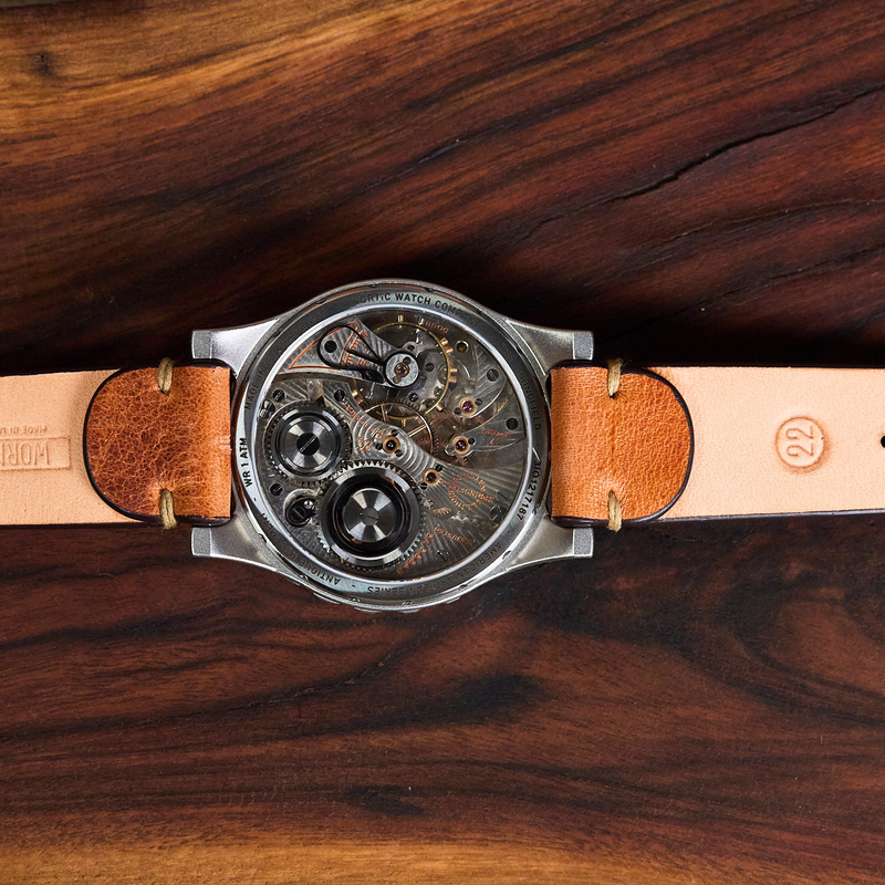 A close-up of a wristwatch with a transparent round case showing a detailed mechanical movement, attached to a light tan leather strap with brown leather ends, laid flat on a dark wood background.