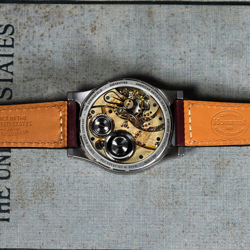 A wristwatch with a transparent back showing a detailed mechanical movement, resting horizontally on a textured gray surface printed with the words 'HISTORIC UNITED STATES' and 'EMERSON FITE'. The watch has a silver-toned case and a tan leather strap with dark brown edges.