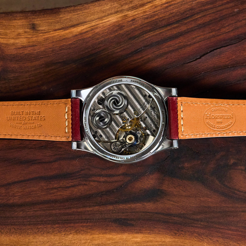 A close-up of a mechanical watch with a transparent case showing the inner movement, paired with a tan leather strap, laid flat on a dark wood background.