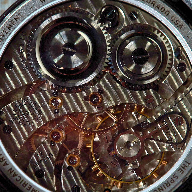 Close-up of a Vostock Watch Company mechanical watch back, showing the intricate silver and gold-toned movement gears and screws, with a black leather strap visible at the edges.