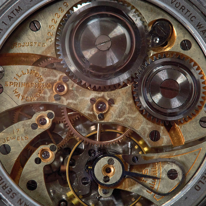 Close-up of a skeleton watch showing an intricate antique movement with gold-toned gears, silver-toned metal parts, and a dark case edge with engraved text.