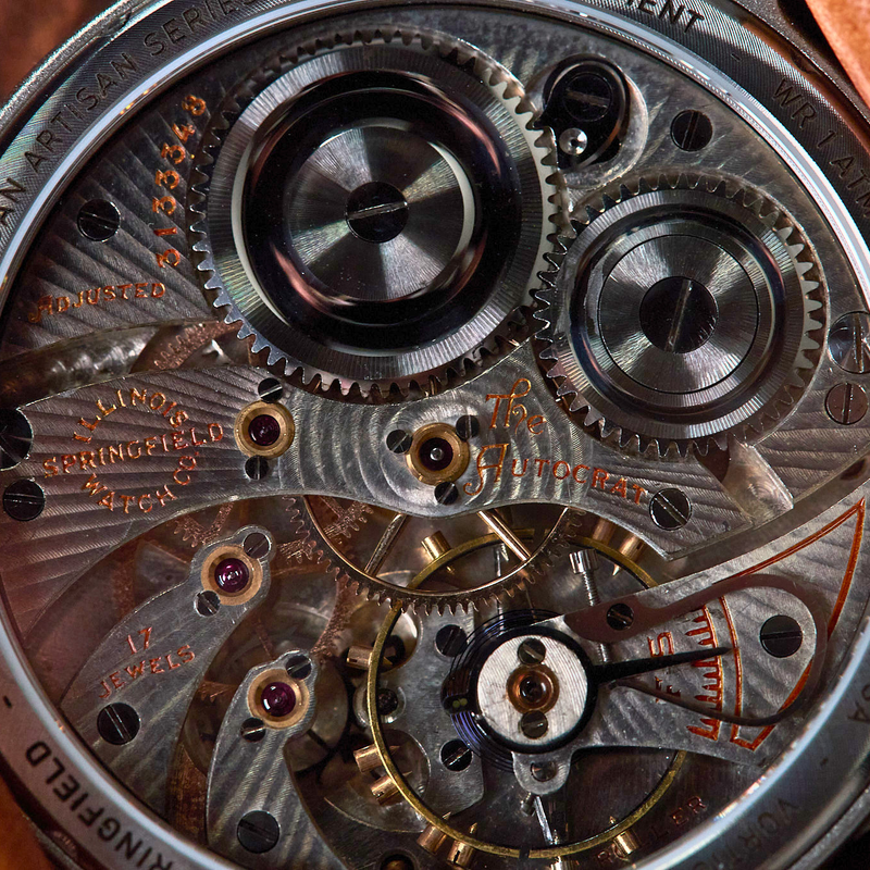 Close-up of a mechanical watch back showing intricate silver and gold-toned gears, screws, and bridges, with engraved text and a brown leather strap edge visible.