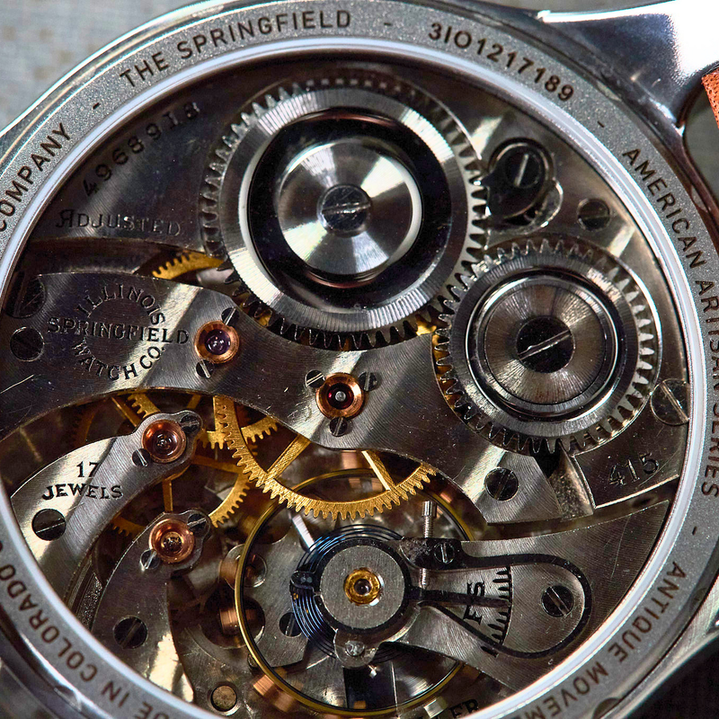 Close-up of a Yortic Watch Company mechanical watch with a silver case, visible internal gears and bridges, and brown leather strap edges on a textured gray background.