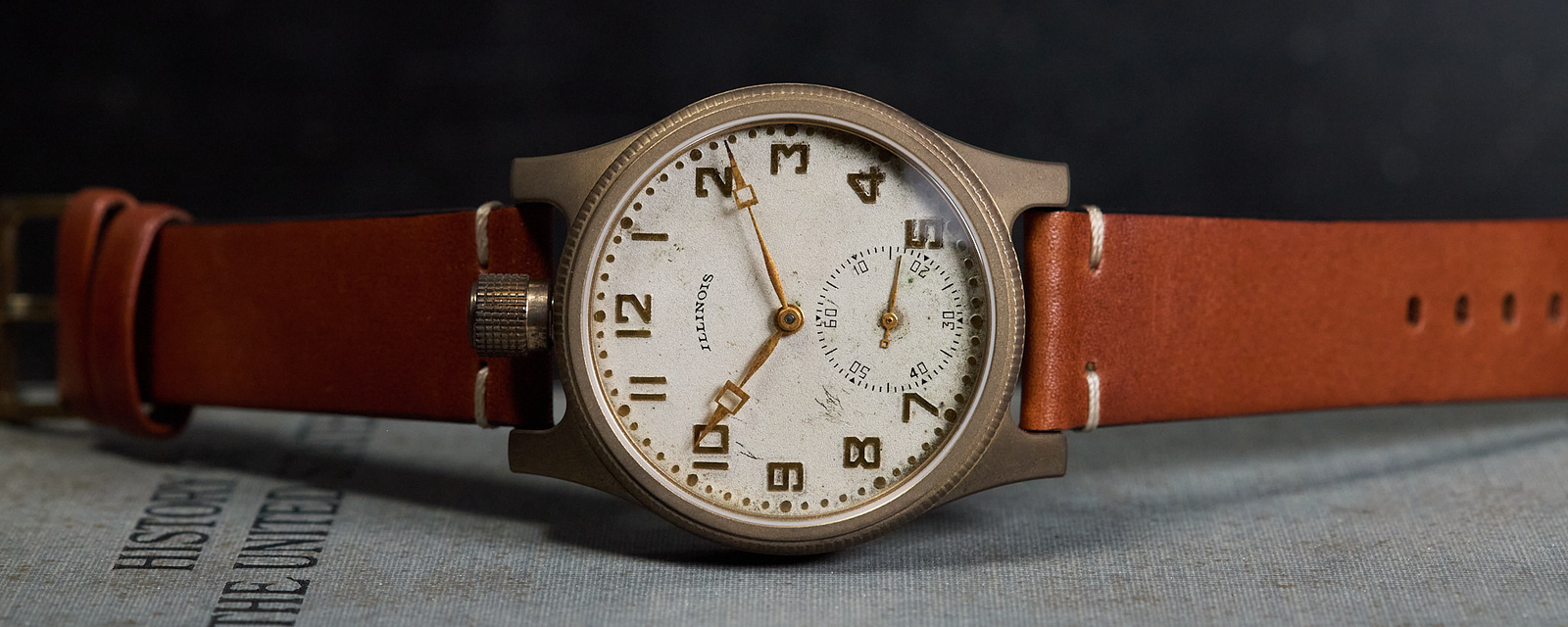 A vintage-style wristwatch with a round bronze-toned case, white dial with black Arabic numerals, and a brown leather strap, resting on a textured gray surface against a dark background.