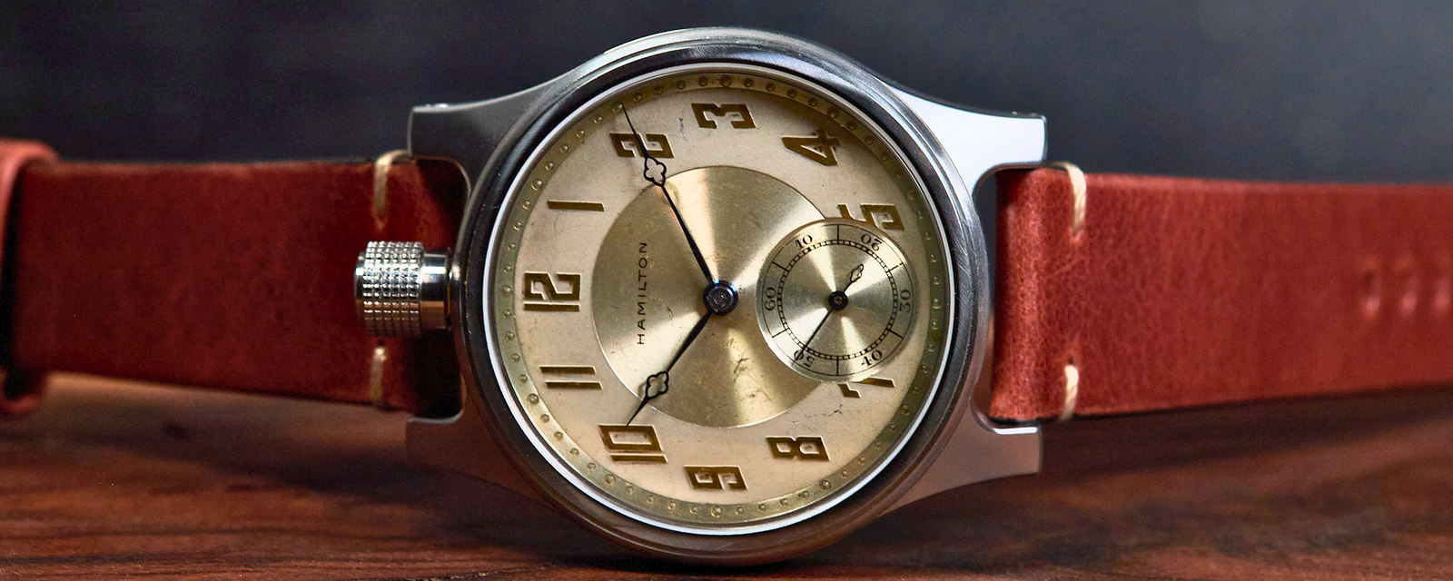 A vintage-style wristwatch with a round silver-tone case, a champagne-gold dial featuring black Arabic numerals and a small subdial, and a reddish-brown leather strap, resting on a wooden surface against a dark background.