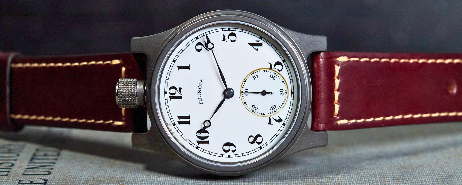 A close-up of a wristwatch with a round silver-toned case, white dial, black Arabic numerals, and a dark red leather strap with light contrast stitching, resting on a textured surface against a dark background.