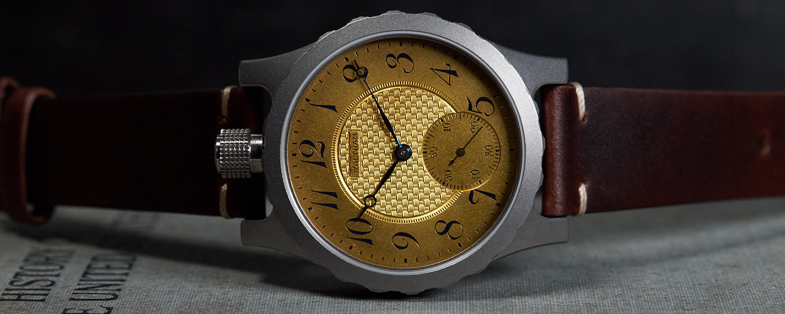 A wristwatch with a textured gold dial, black Arabic numerals, a silver-toned case, and a dark brown leather strap, resting on a gray textured surface against a dark background.