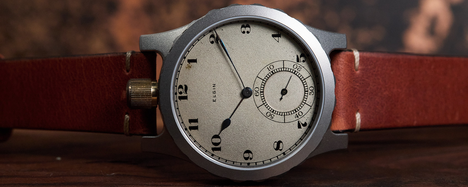A vintage-style wristwatch with a round silver-toned case, cream-colored dial, black Arabic numerals, and a brown leather strap, resting on a wooden surface with a blurred rustic background.