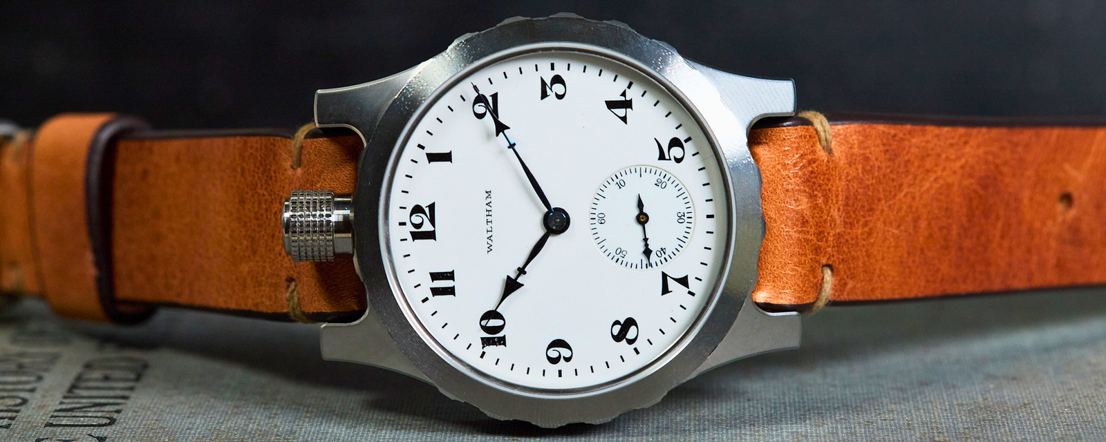 A wristwatch with a silver-toned metal case, a white dial with black Arabic numerals, and a brown leather strap, displayed on a textured gray surface against a dark background.