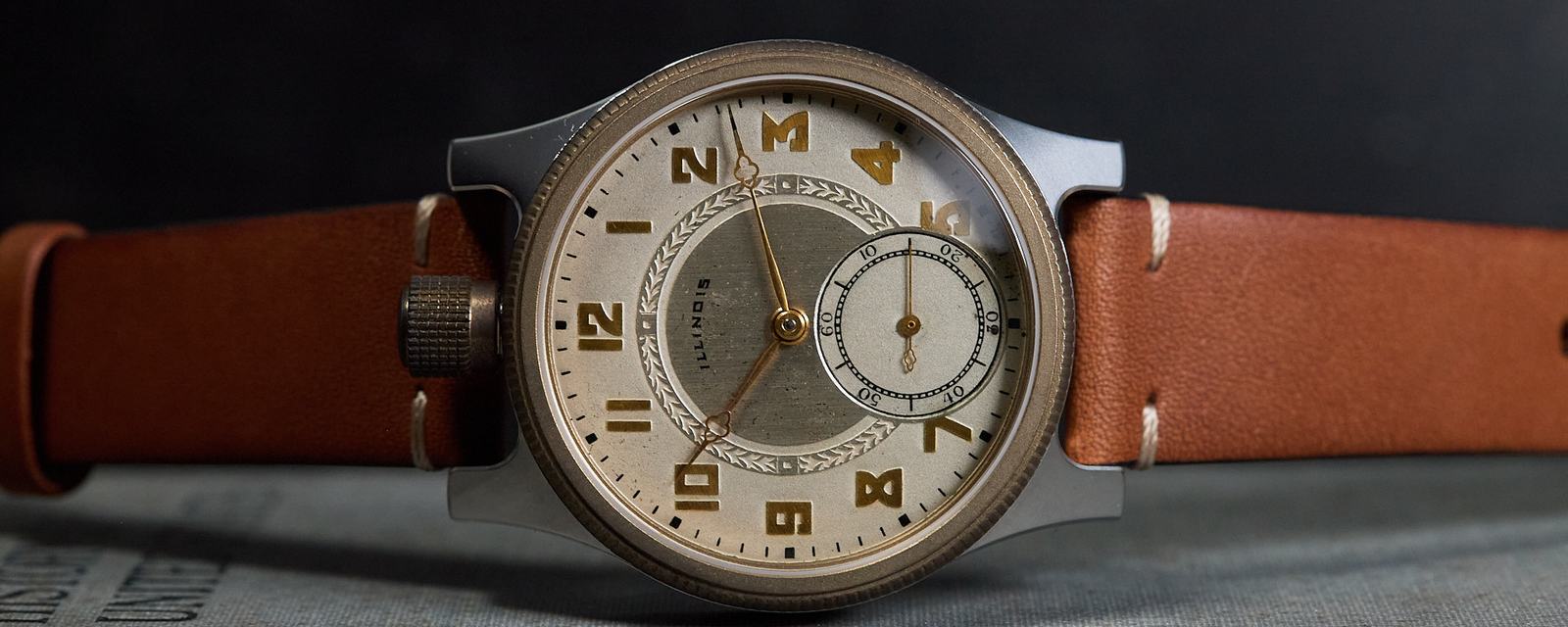 A vintage-style wristwatch with a round silver-toned case, cream dial, brown leather strap, and gold-tone numerals, resting on a textured gray surface against a dark background.