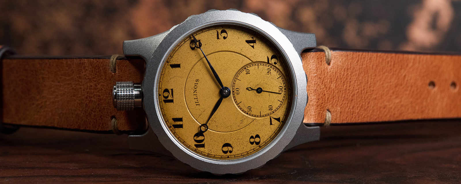 A wristwatch with a round silver-tone case, a warm tan dial with black Arabic numerals and hands, and a brown leather strap, photographed on a dark wooden surface with a blurred rustic brown background.